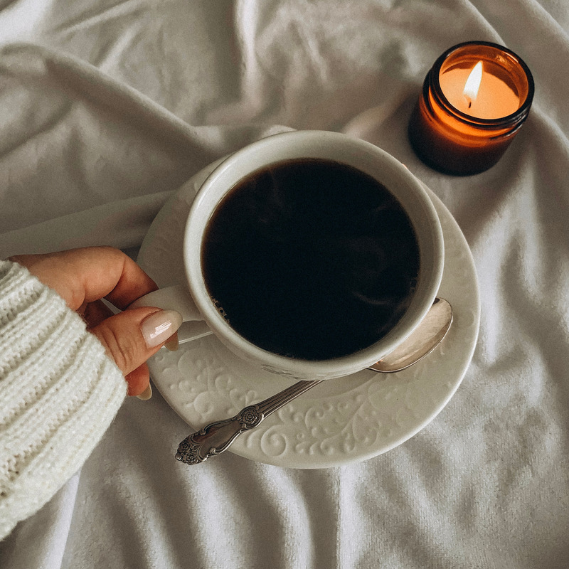 A coffee cup on a saucer beside a lit candle on a soft white blanket